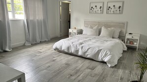 A spacious bedroom with a large bed, white bedding, and a wooden floor. There are two bedside tables with lamps, a nightstand, and a potted plant. - Walk to the Beach and everything else (Hollywood)