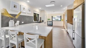A modern kitchen with a central island, white chairs, wooden cabinets, and a view to the outdoors. - Sorrento Oasis - 5BR, Gym, Pool (Bundall)