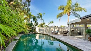 A swimming pool with a clear blue water, surrounded by palm trees and a patio area with outdoor furniture. - Sorrento Oasis - 5BR, Gym, Pool (Bundall)