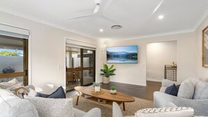 A modern living room with a large window, a ceiling fan, a wooden coffee table, and a sofa. - Sorrento Oasis - 5BR, Gym, Pool (Bundall)