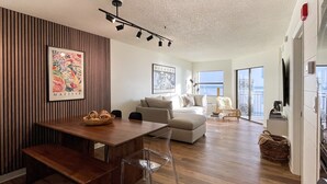 Una sala de estar moderna con una mesa de comedor de madera, sillas traslúcidas, un sofá y un ventanal grande.