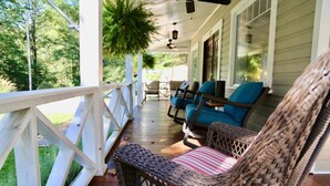 A covered porch with wicker chairs and a table, surrounded by white railings and greenery.