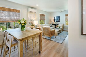 A spacious living area with a wooden dining table, wicker chairs, a sofa, and a coffee table. There are two windows with bamboo blinds, an air conditioning unit, and a ceiling fan.
