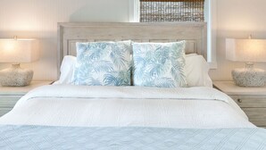 Een net opgemaakt bed met een lichtblauwe quilt, twee bijpassende kussens met een tropisch bladmotief, en twee ronde nachtkastjes met lampen en decoratieve vazen.