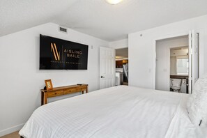 Une chambre à coucher avec un grand lit, une commode en bois, un téléviseur fixé au mur et une porte donnant sur une salle de bain.