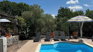 Uma piscina com espreguiçadeiras e área coberta, cercada por árvores e um muro de pedra.