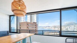Un balcon avec une table en bois, deux tabourets et un banc. Une grande fenêtre offre une vue sur le paysage urbain et sur une étendue d’eau.