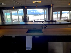 A kitchen with a large window overlooking the beach, a sink, and hanging pendant lights.