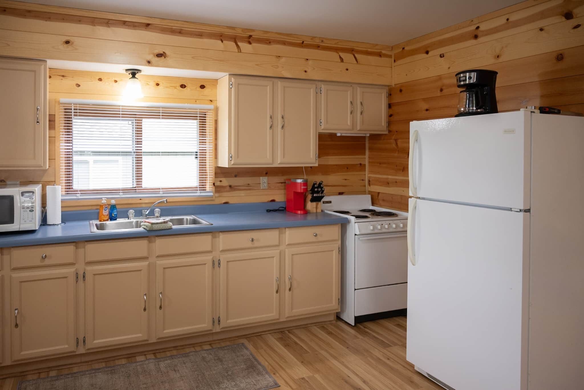 Une cuisine avec des murs en bois, un réfrigérateur blanc, une cuisinière, un four à micro-ondes et un évier sur lequel se trouve une fenêtre.