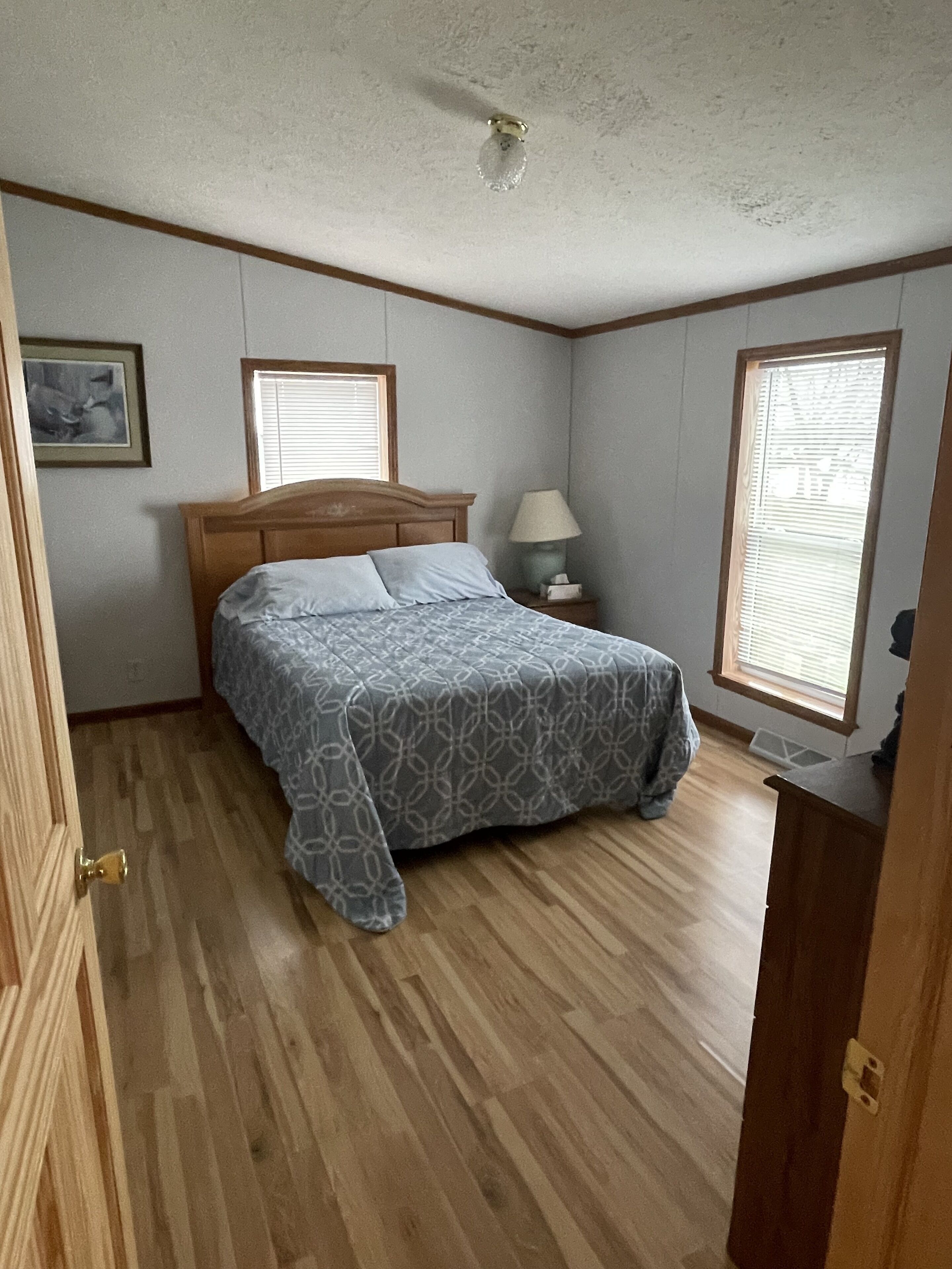A bedroom with a wooden bed, two windows with blinds, a bedside lamp, and a framed picture on the wall.