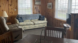 Ein gemütliches Zimmer mit Holzwänden, einem Sofa mit floralem Bezug, einem Holzstuhl und einem Esstisch.