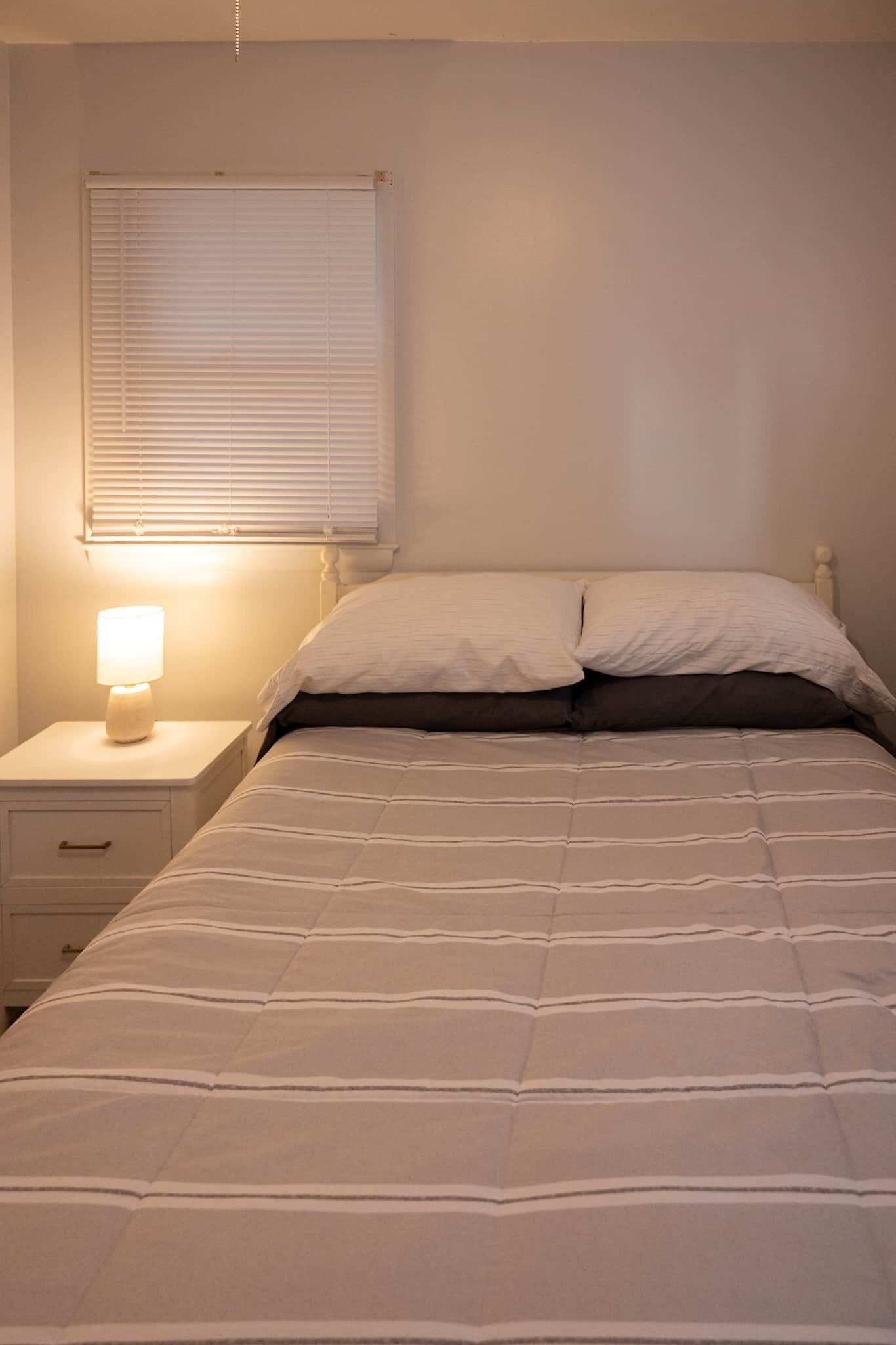 A bedroom with a bed, a nightstand with a lamp, and a window with blinds.