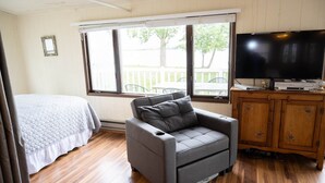 Ein Zimmer mit einem Bett, einem grauen Sofa, einem Fernseher auf einer Holzkommode und einem Fenster mit Blick auf eine Terrasse und Bäume.