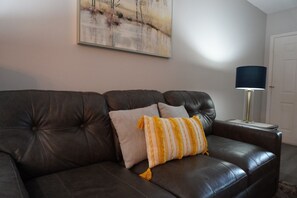 En mørk lædersofa med to puder, en lampe og et maleri på væggen.