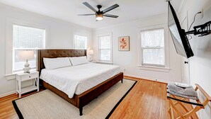 Une chambre à coucher comprenant un lit, une table de chevet, une lampe, un ventilateur de plafond et un téléviseur fixé au mur.