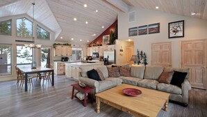 Un vaste espace de vie doté d’un haut plafond, d’un plancher en bois, d’un coin salon central avec un canapé et une table basse, et d’une cuisine à l’arrière-plan.