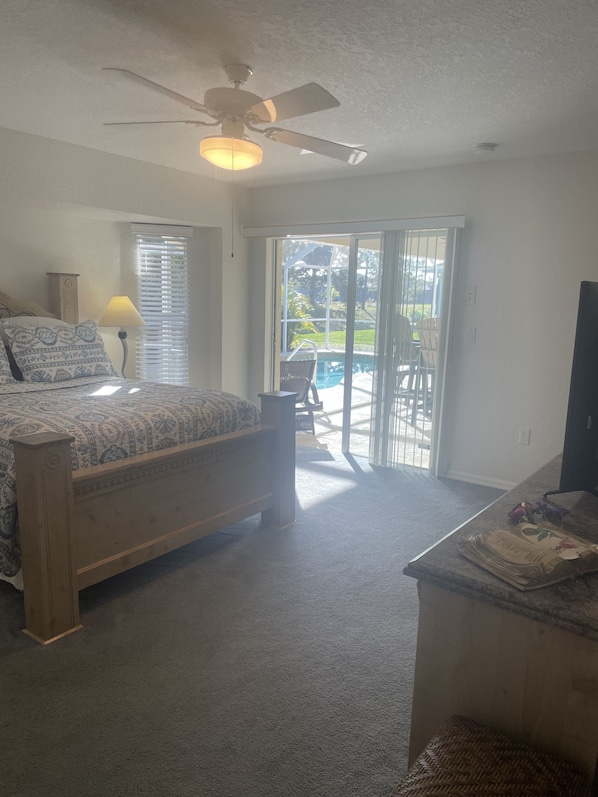 A bedroom with a bed, a ceiling fan, a lamp, and a view of a patio with a pool.