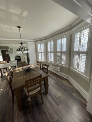 A spacious dining area with a wooden table and chairs, large windows with shutters, and a kitchen in the background.