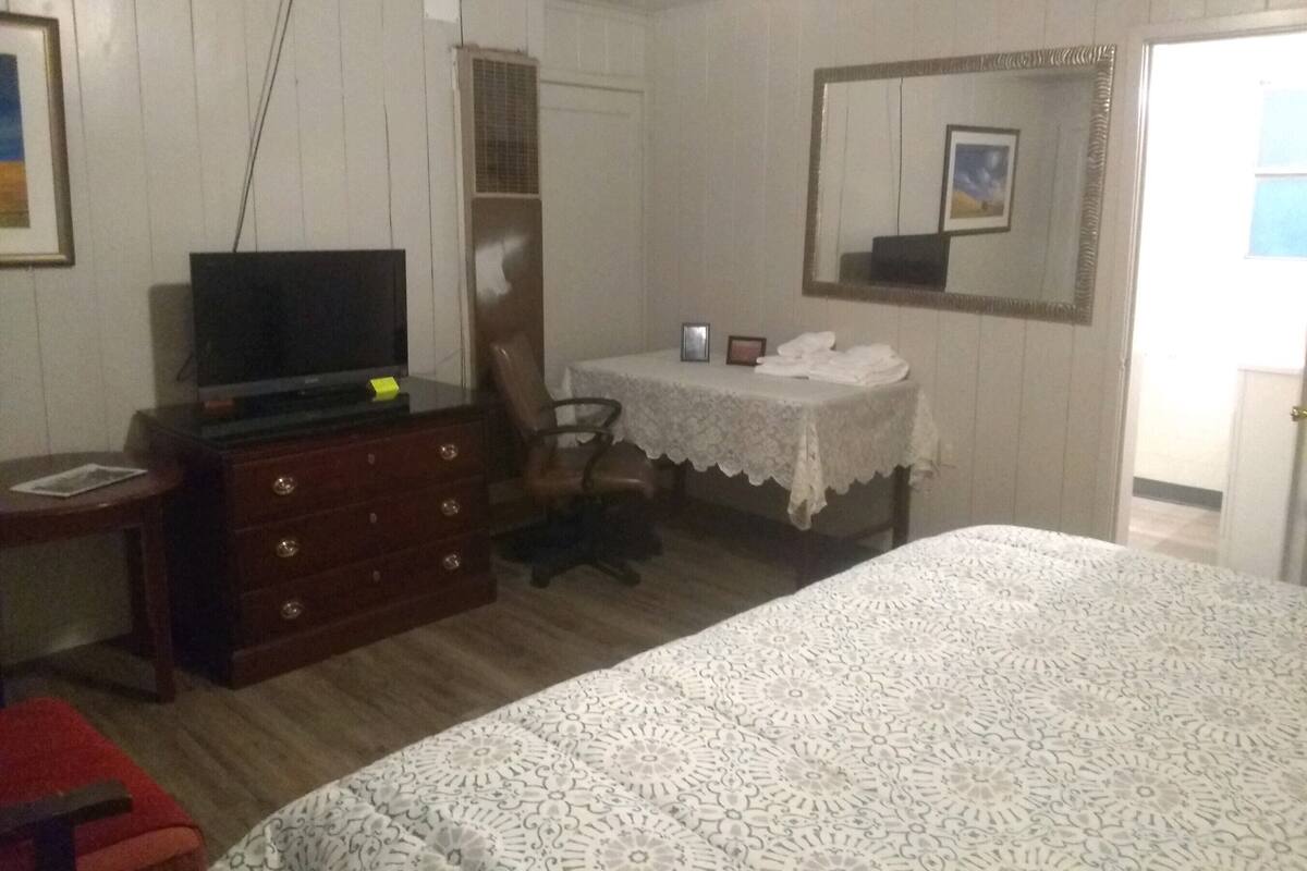 Ein Schlafzimmer mit Bett, Schminktisch, Fernseher, Schreibtisch mit Stuhl, Spiegel und einer Tür.