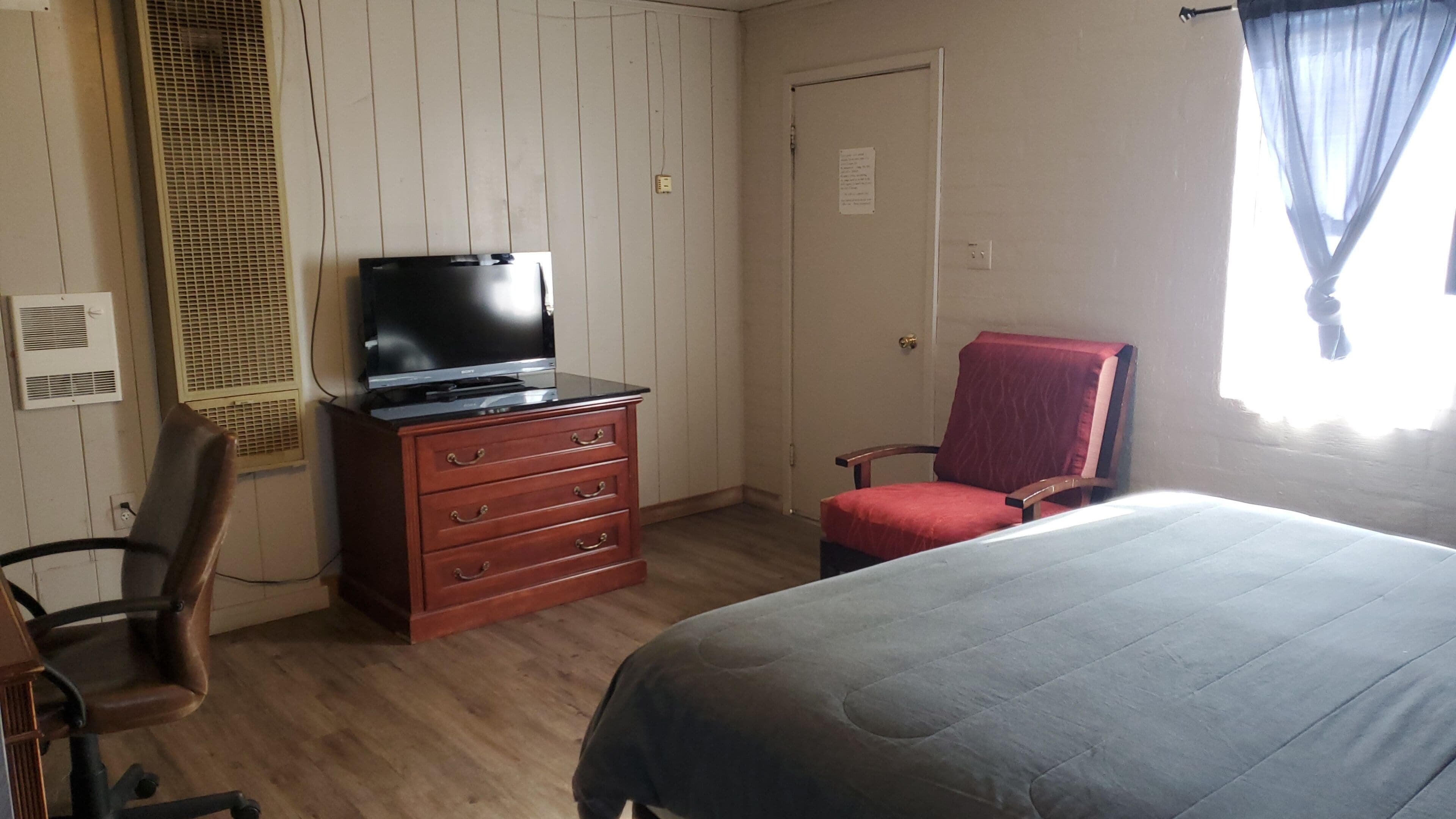 Une chambre à coucher avec un lit, un téléviseur posé sur une commode, une chaise et une fenêtre avec des rideaux.