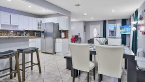A modern kitchen with white cabinets, a stainless steel refrigerator, and a dining area with a table and chairs. - Posturepedic Beds!NearBeach/Airport Spacious BBQ/BAR (Miami)