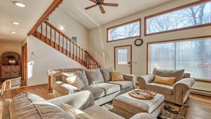 Ein geräumiges Wohnzimmer mit hoher Decke, einer Holztreppe und großen Fenstern mit Blick auf Bäume.