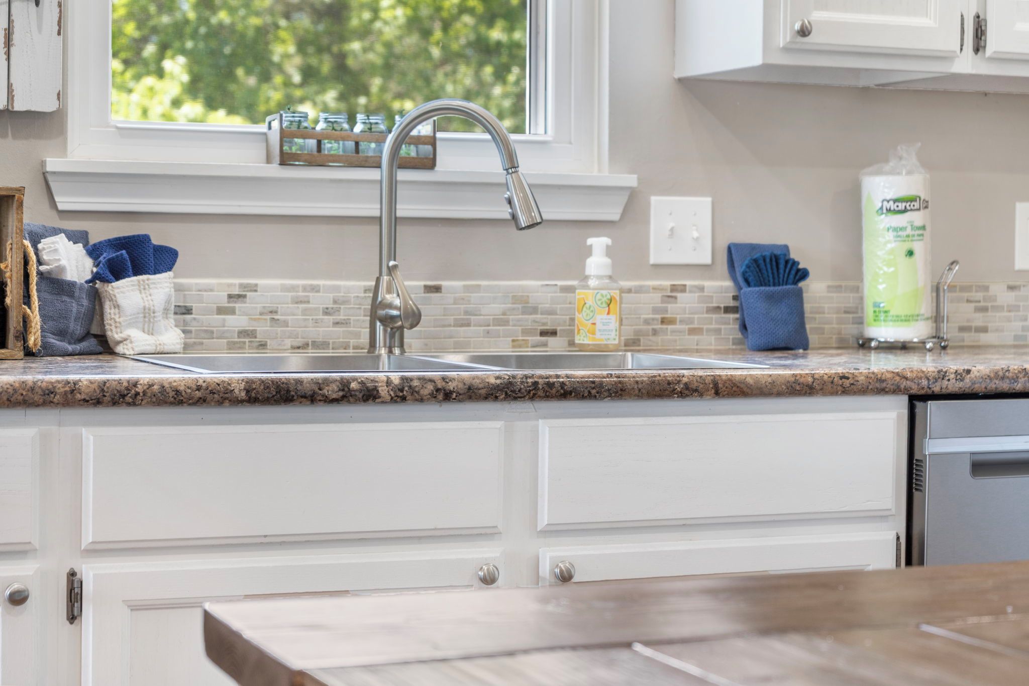 A kitchen with a stainless steel sink, a window with a view of greenery, and various kitchen items on the countertop.