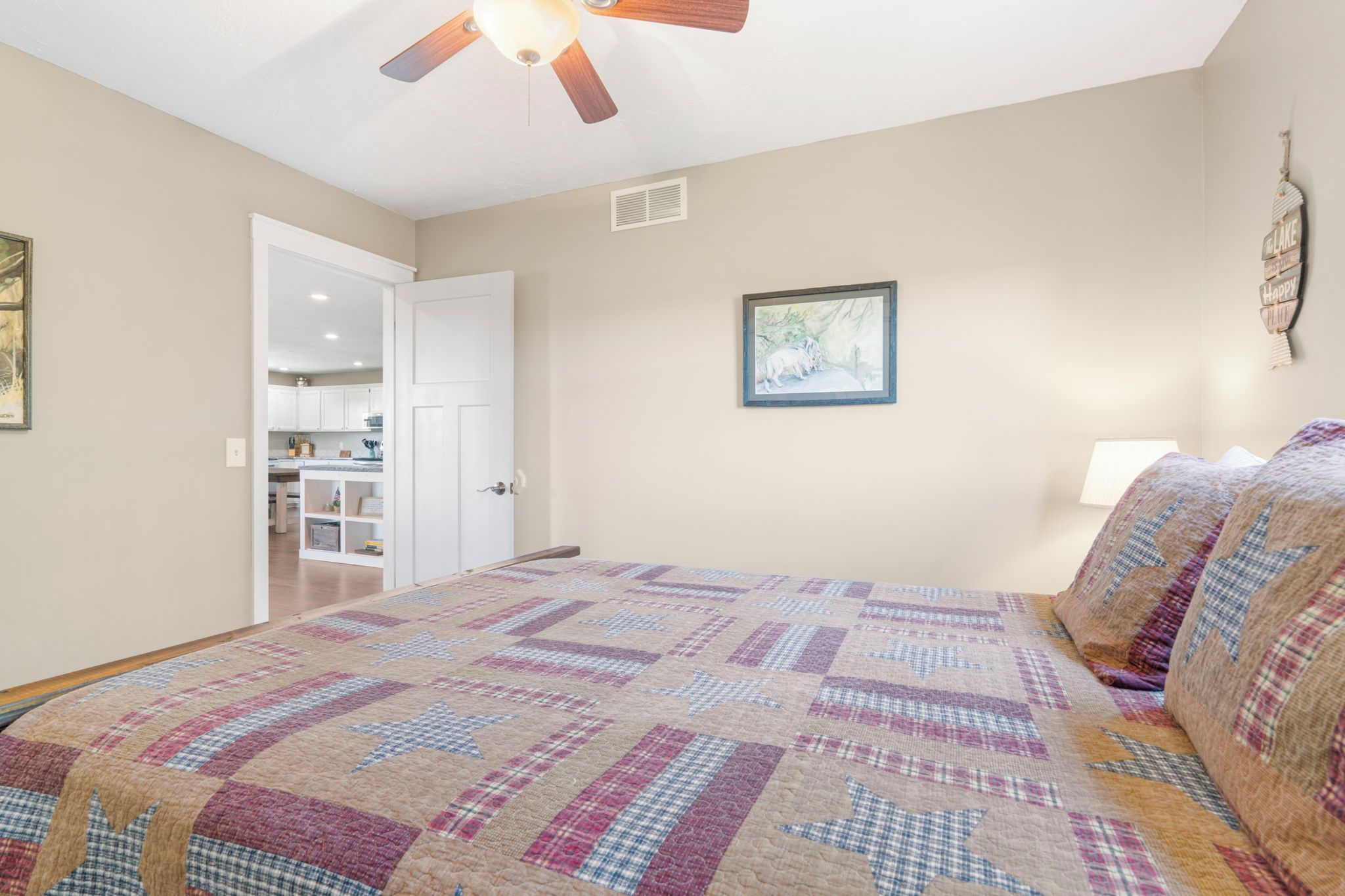 A bedroom with a bed, a ceiling fan, a door leading to a kitchen, and a wall-mounted picture.