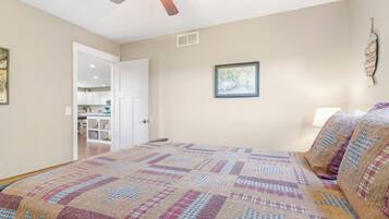 A bedroom with a bed, a ceiling fan, a door leading to a kitchen, and a wall-mounted picture.
