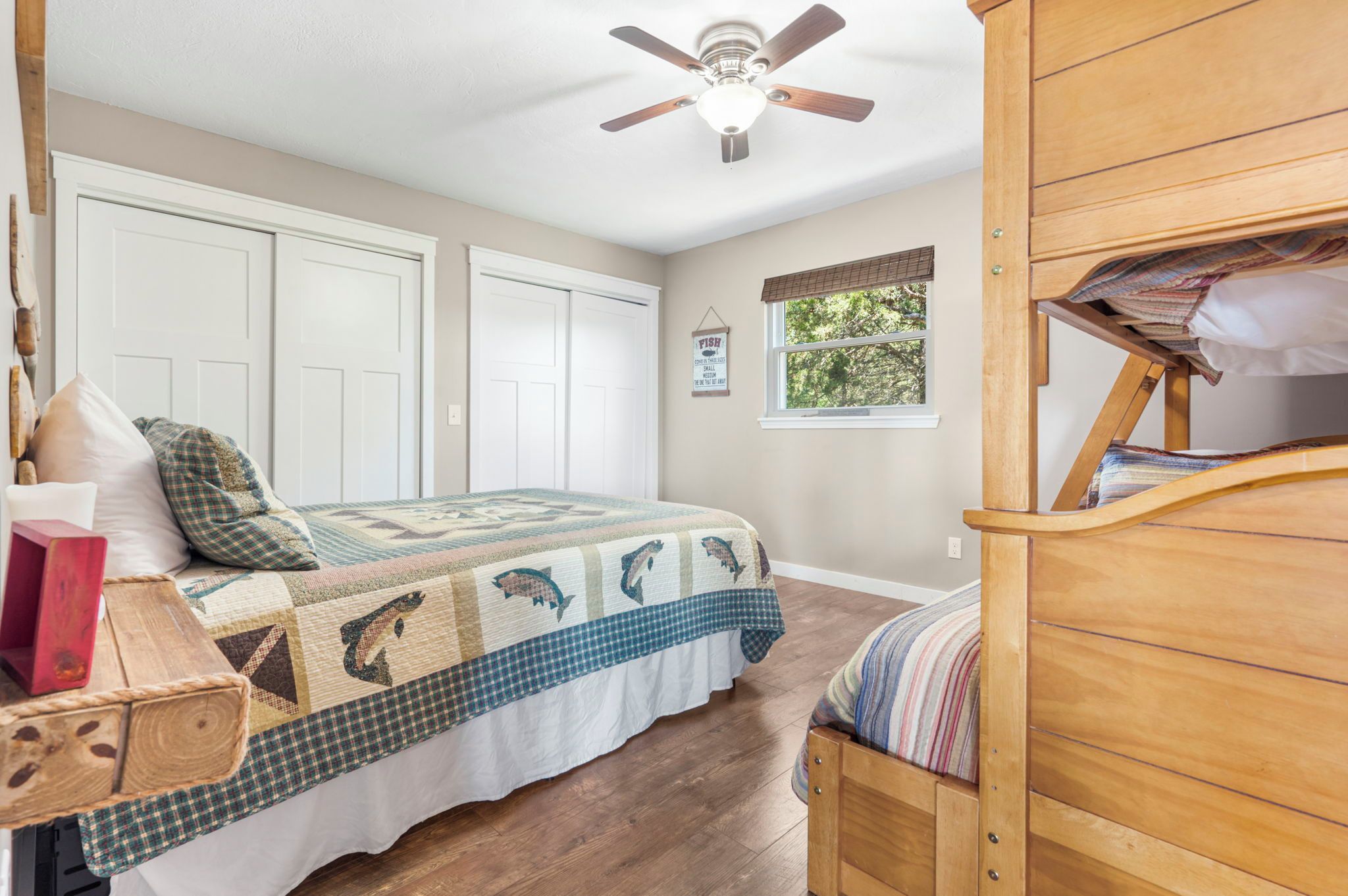 A bedroom with a bed, a wooden dresser, a ceiling fan, and a window with a view of trees.