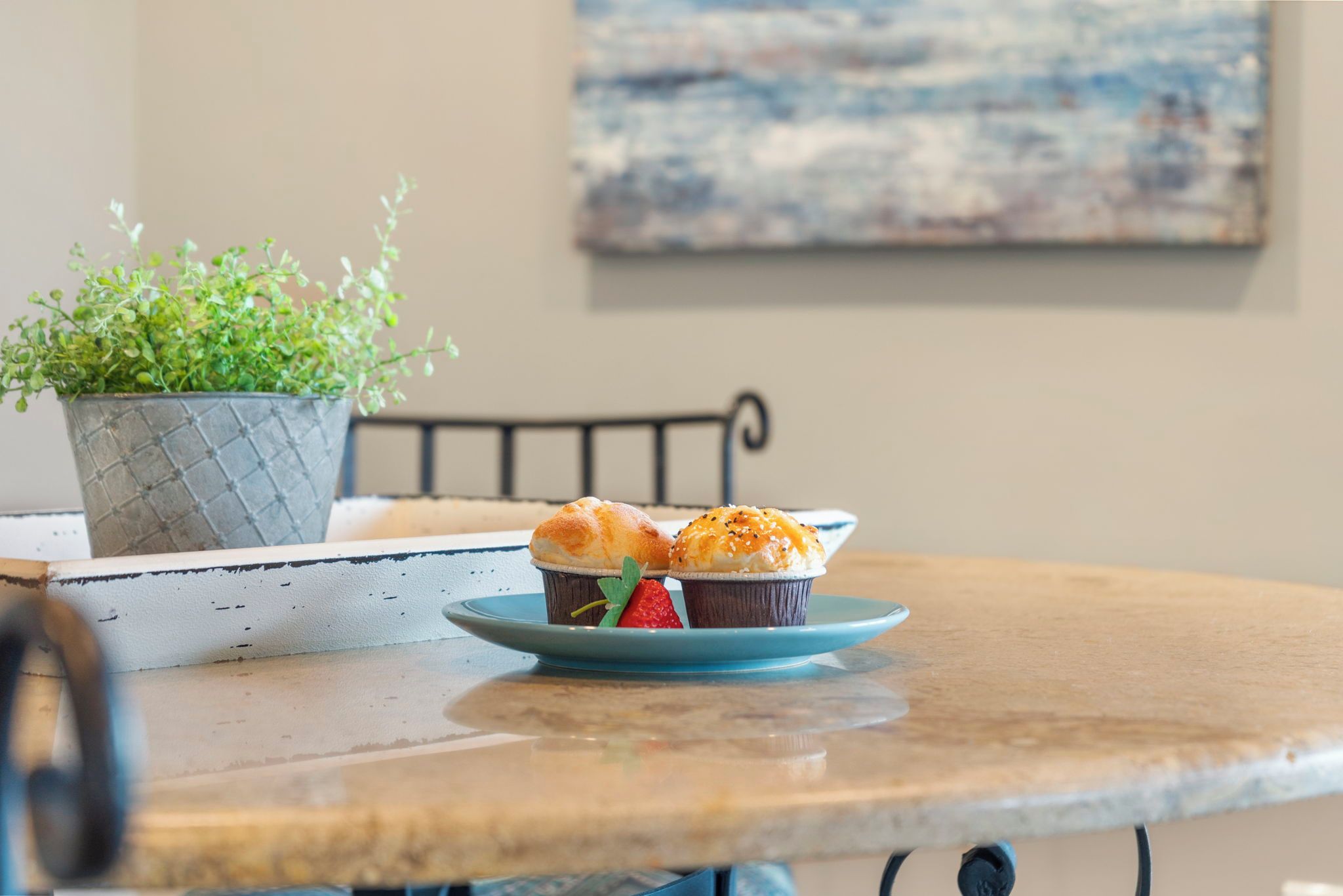 A round table with a blue plate holding two muffins and a strawberry, a potted plant, and a metal chair.