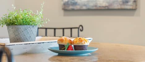Una mesa redonda con un plato azul que contiene dos muffins y una fresa, una planta en maceta y una silla metálica.