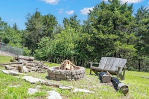 A fire pit surrounded by logs and rocks, a wooden bench, and a chain-link fence in a wooded area. - Secluded Getaway featuring Walking Trail to the Lake, Free Tickets & Minutes from Silver Dollar City (Reeds Spring)