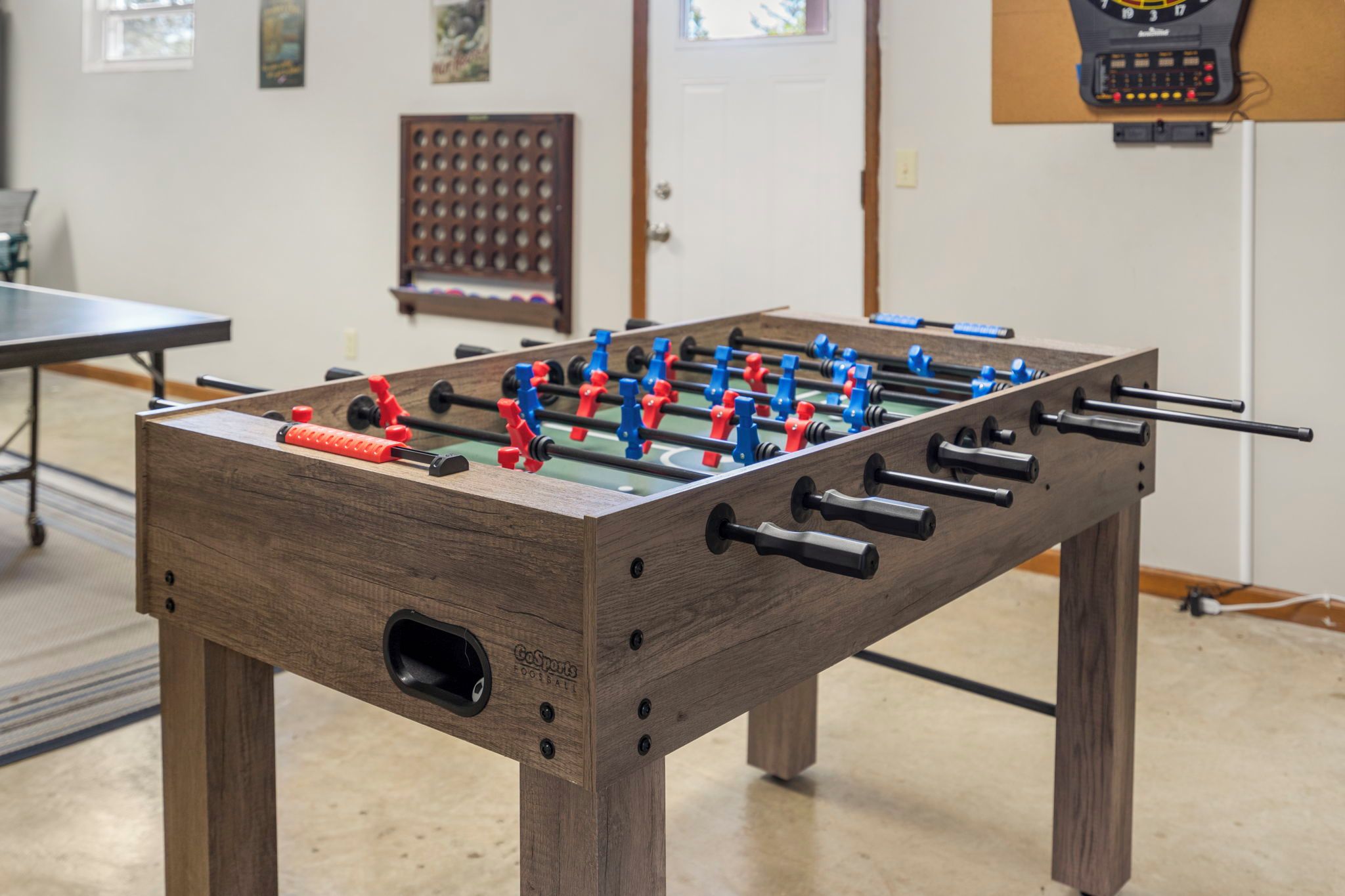 A wooden foosball table with red and blue players, set in a room with a ping pong table and a scoreboard in the background.