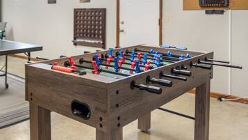 A wooden foosball table with red and blue players, set in a room with a ping pong table and a scoreboard in the background.