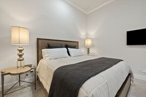 Un dormitorio ordenado con cama, lámparas de noche, una mesita y un televisor de pantalla plana en la pared.