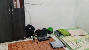 Um quarto pequeno com uma cama, uma escrivaninha com um laptop, um capacete verde e uma bolsa preta.