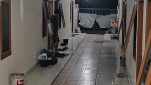 Um corredor com piso de azulejos, uma pessoa próxima a uma parede e diversos objetos espalhados pelo chão.