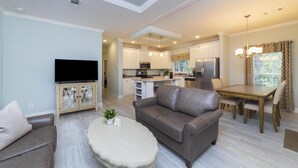 Un soggiorno moderno con un divano in pelle, un tavolino da caffè e un televisore su un mobile. Si intravede una zona pranzo con un tavolo e sedie di legno. La cucina è dotata di elettrodomestici moderni.