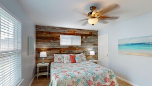 Une chambre avec un lambris en bois, un lit recouvert d’une literie à motifs floraux, un ventilateur de plafond et un tableau représentant une plage.