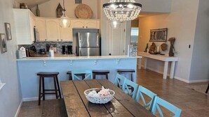 Une cuisine spacieuse dotée d’un îlot central, un coin repas avec une table en bois et des chaises bleues, ainsi qu’un lustre.