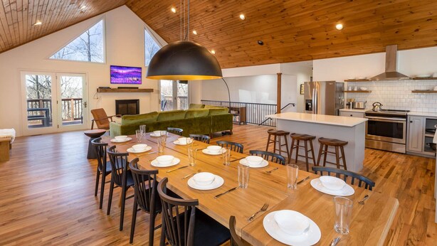 Un vaste espace de salle à manger avec une longue table en bois dressée pour le repas, un confortable canapé vert et une cuisine équipée d’appareils modernes.