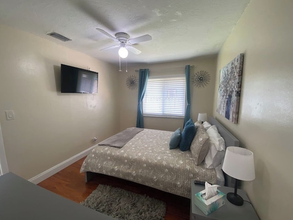 A bedroom with a bed, a ceiling fan, a TV mounted on the wall, and a window with curtains.