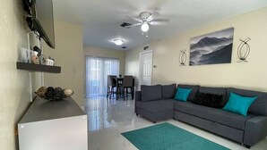 A living room with a grey sectional sofa, a teal rug, a ceiling fan, and a dining area with a table and chairs.
