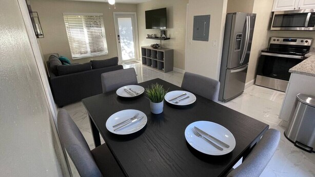 A modern kitchen with stainless steel appliances, a dining area with a dark wooden table set for four, and a living area with a grey sofa.