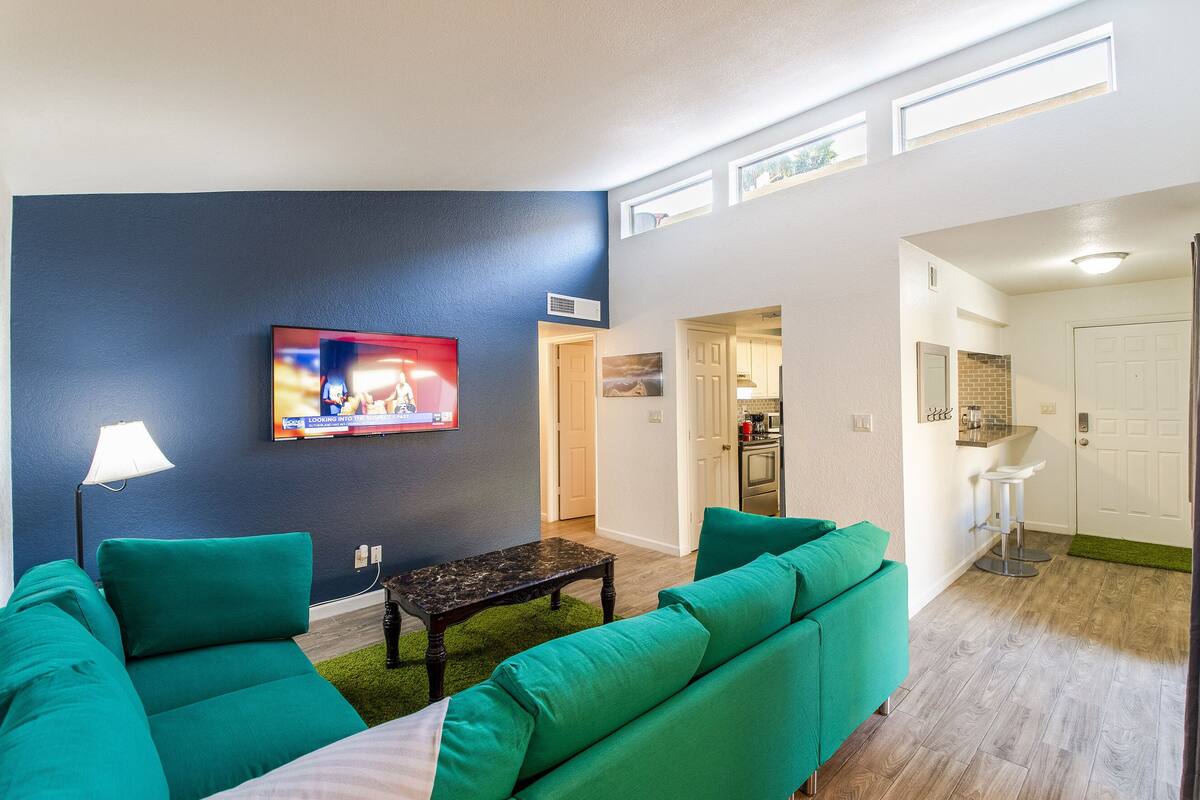 A living room with teal sofas, a TV on the wall, a small coffee table, and a kitchen area visible through an open door.