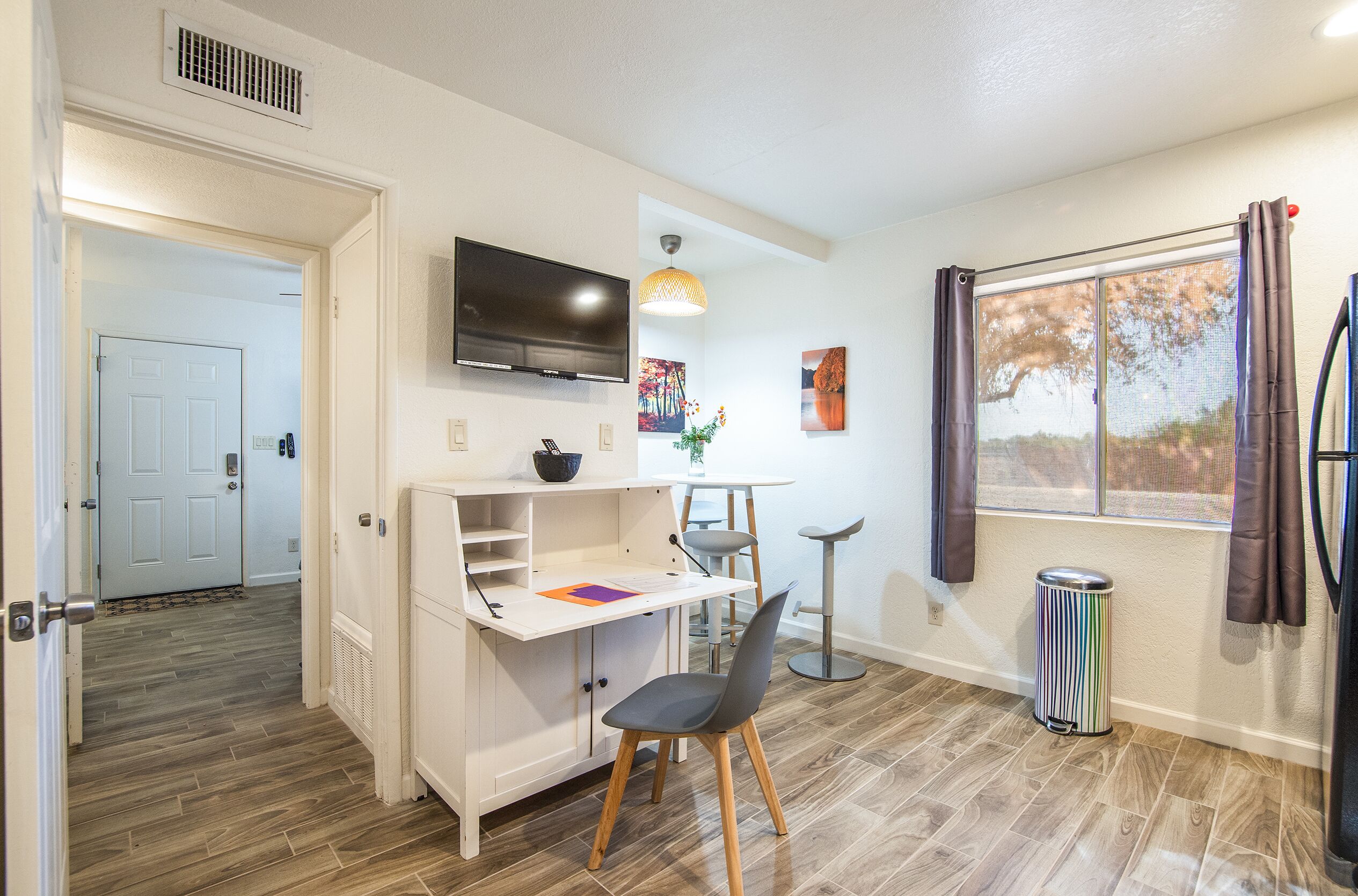 Un espace de vie compact comprenant un coin cuisine, un téléviseur fixé au mur, un coin repas avec une table et des chaises, et une fenêtre avec des rideaux.