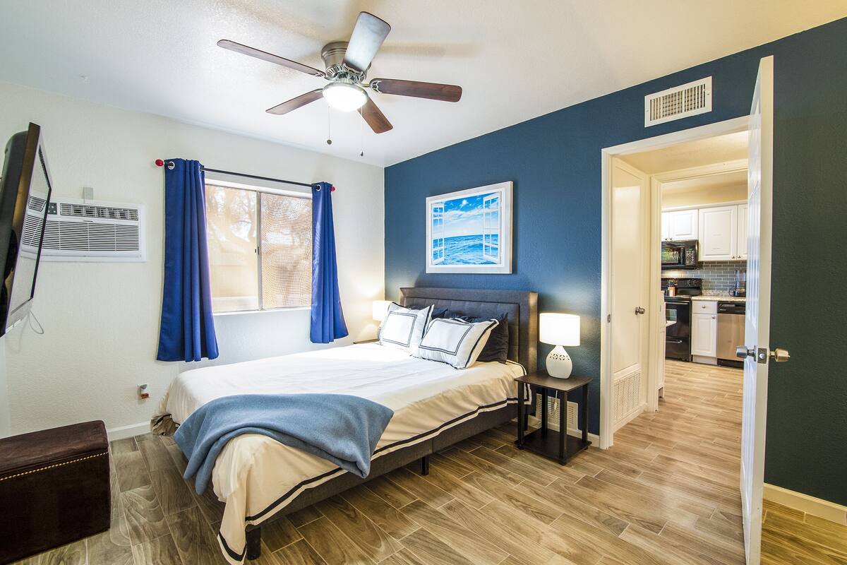 A bedroom with a bed, a ceiling fan, a window with curtains, a bedside table with a lamp, and a view into the kitchen.
