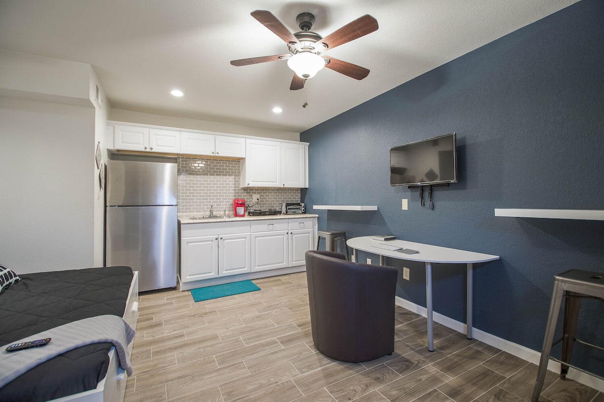 A compact kitchen with white cabinets, a stainless steel refrigerator, a built-in oven, a microwave, and a coffee maker. There's a dining area with a round table and a single chair, and a ceiling fan above.