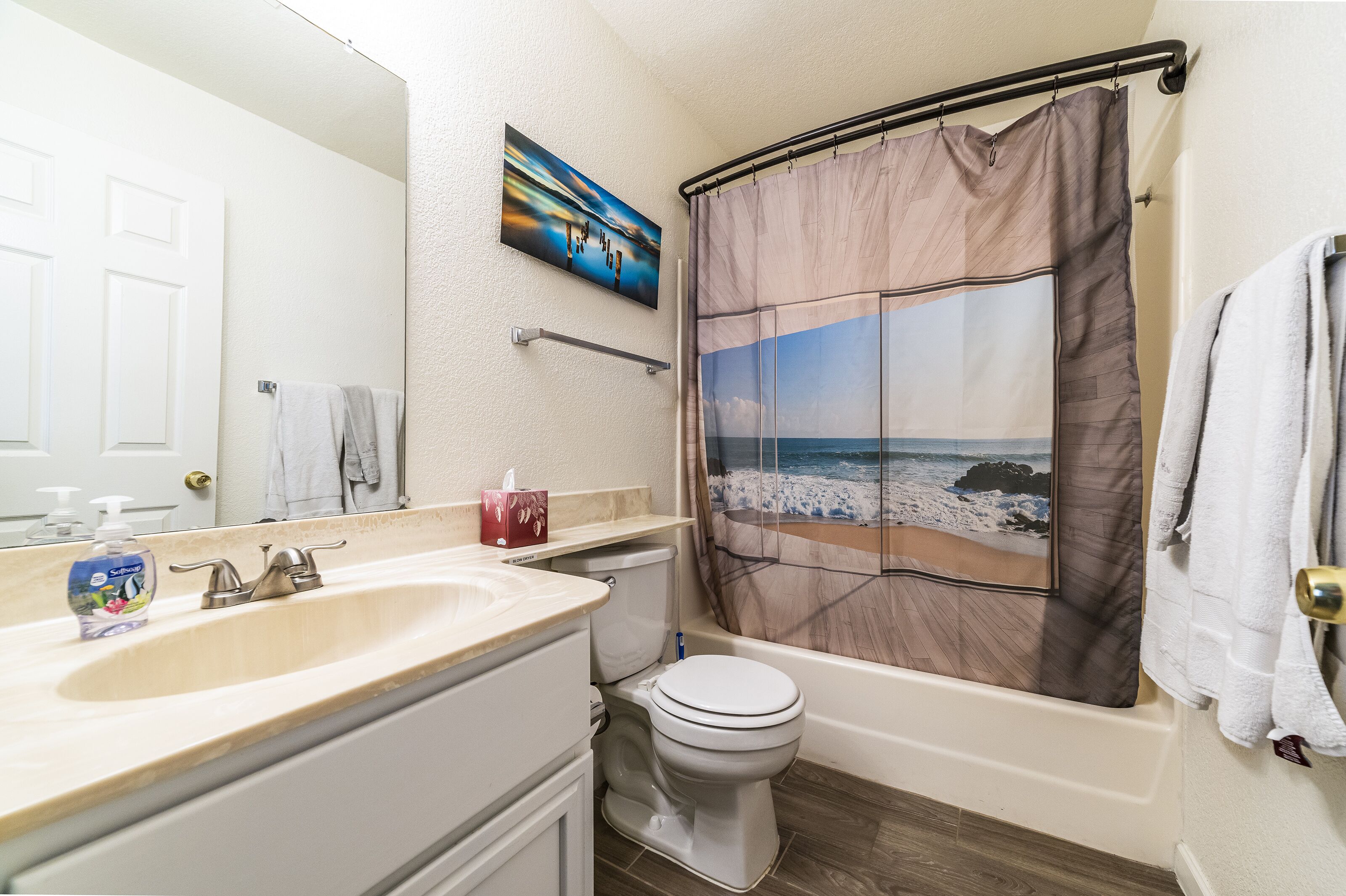 A bathroom with a beige sink, a white toilet, a shower curtain with a beach scene, and a towel rack with a white towel.
